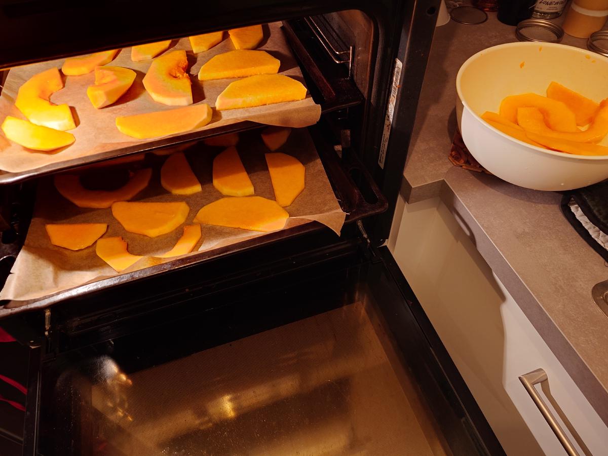 Two parchment-lined trays of squash going into the oven, a bowl of leftover slices on the counter