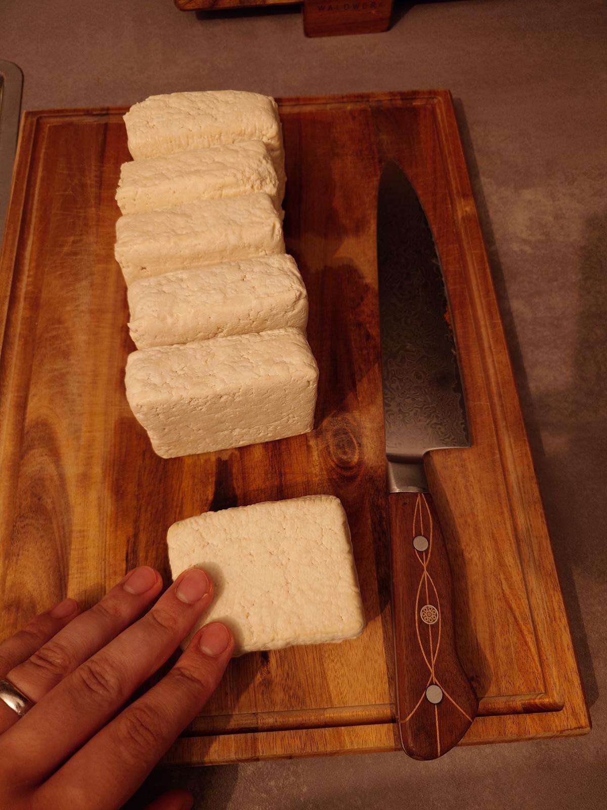 Six pressed slabs of tofu leaning in a row on a wooden board beside a knife
