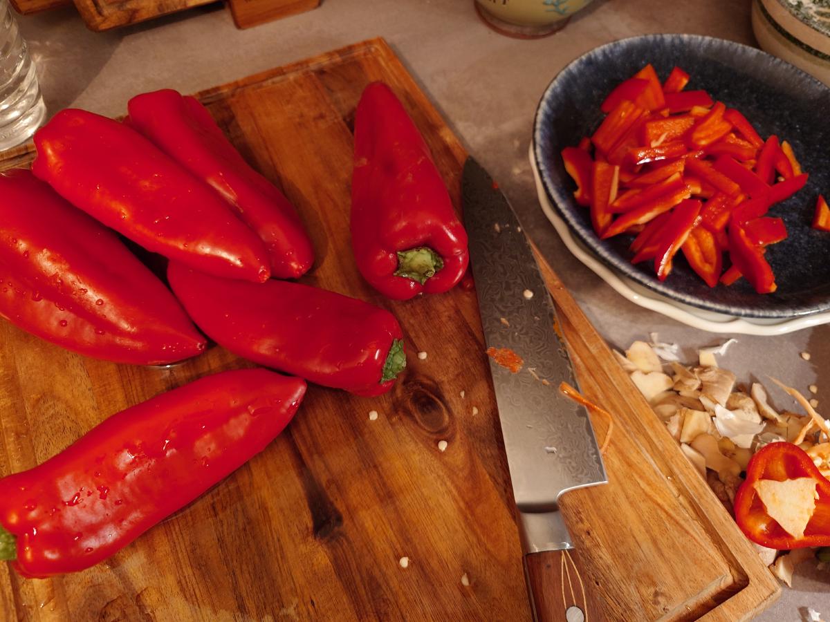Pointed red peppers on the board, six still whole, one already cut into strips in a dark bowl
