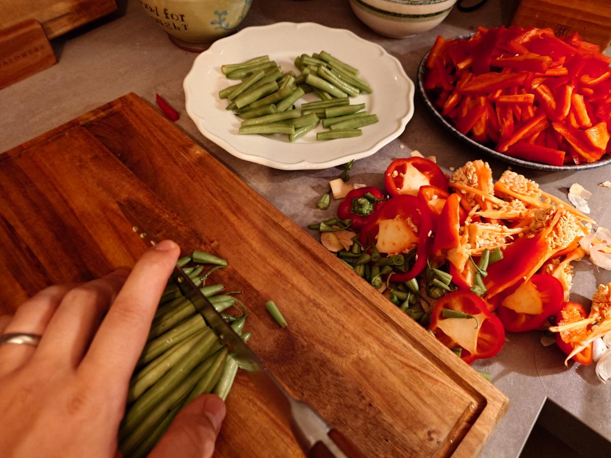 Green beans cut in half on the board, a plate of cut beans and a bowl of pepper strips behind