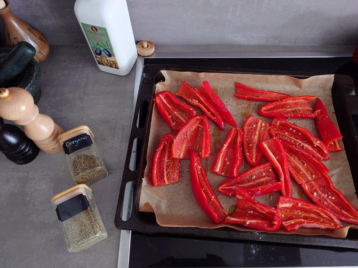Red pepper strips spread on a parchment-lined tray, oregano and rosemary jars and the olive oil bottle beside it