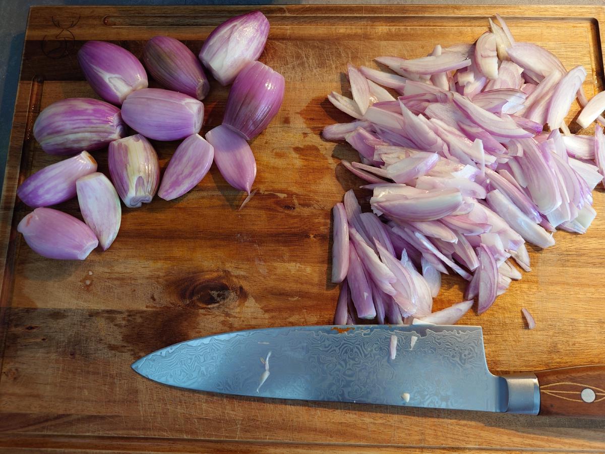 The board of shallot chunks beside the whole ones still to cut, the knife along the bottom