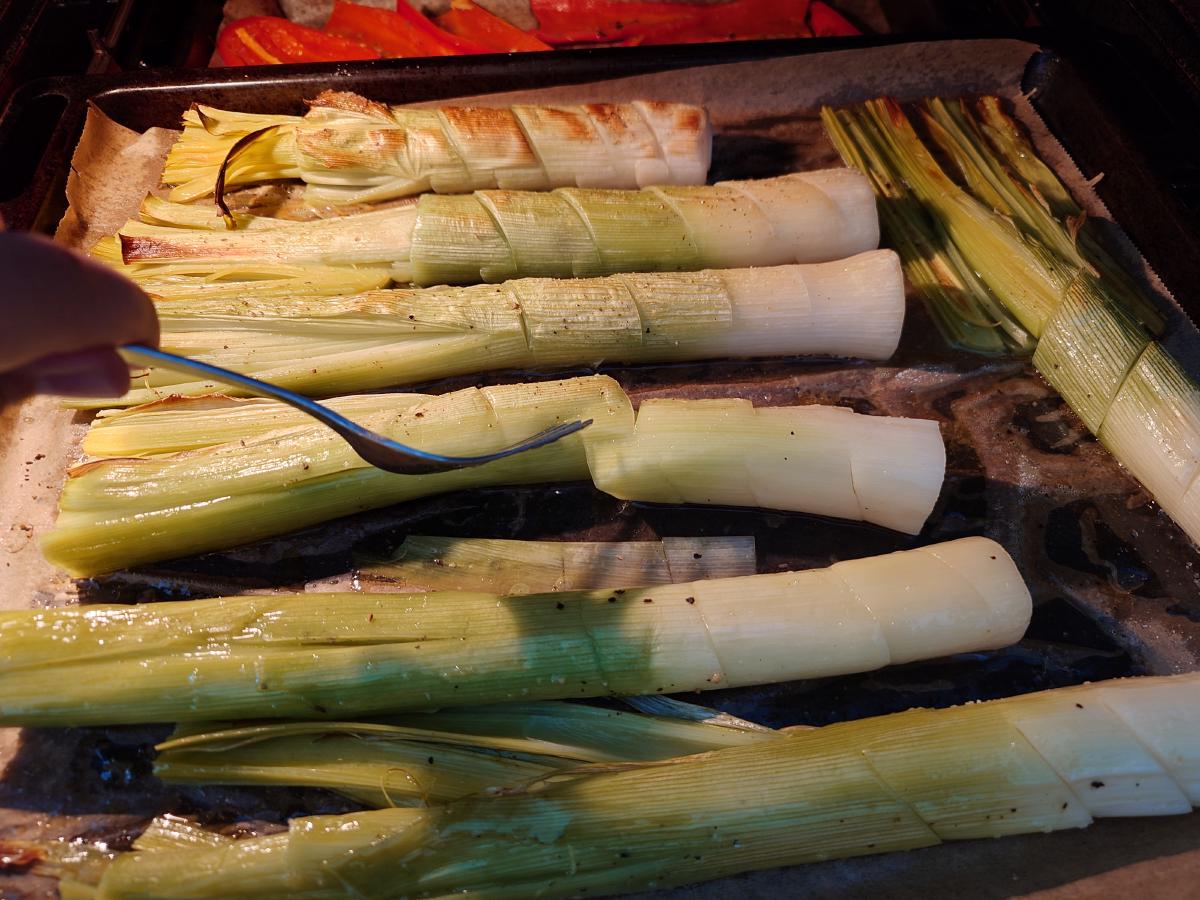 A fork under a leek, turning it over on the tray in the oven, the peppers browning on the shelf above