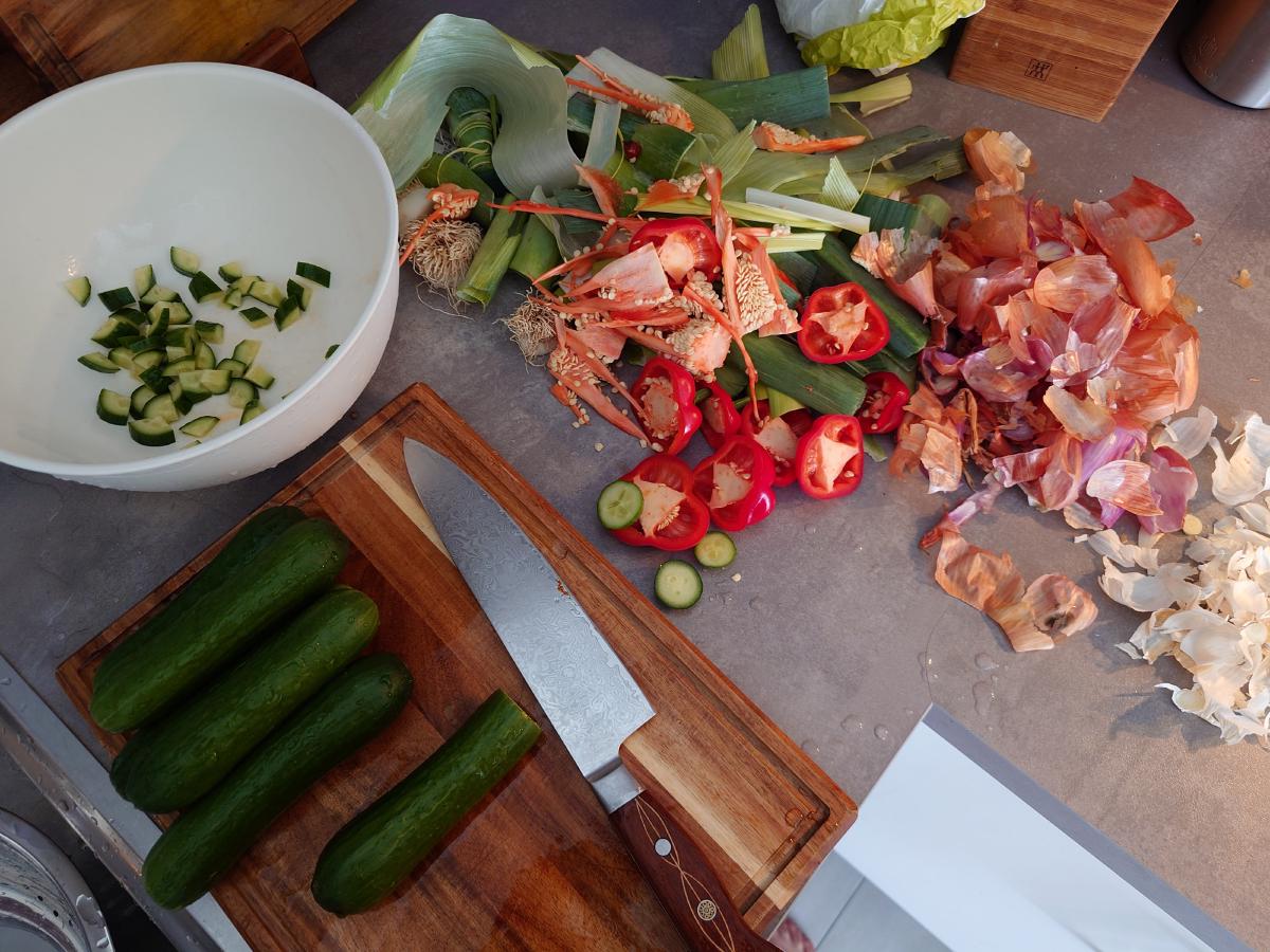 Four cucumbers on the board, cut chunks already in the white bowl, a long pile of leek, pepper and shallot trimmings across the counter behind