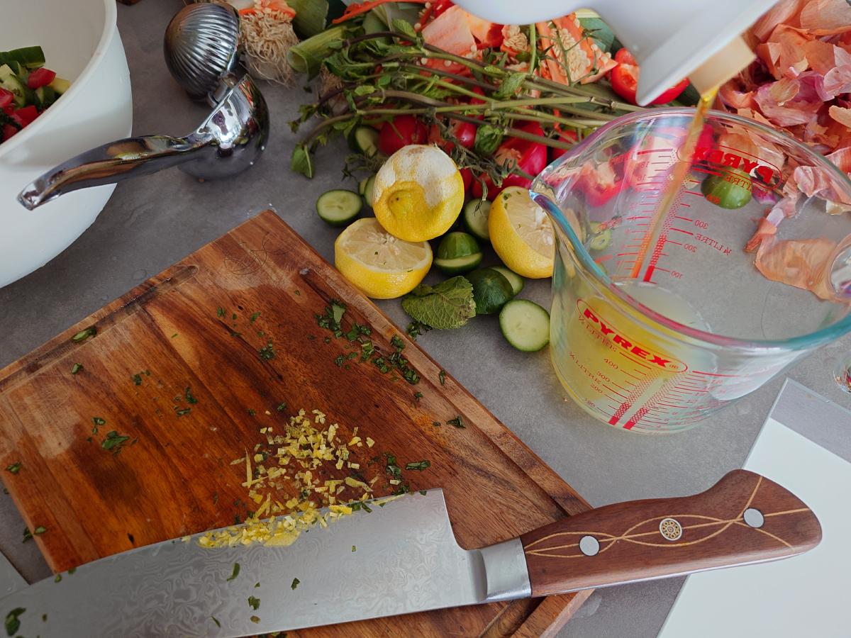 Lemon zest and chopped herbs on the board, squeezed lemon halves and the press behind, olive oil pouring into a jug of lemon juice