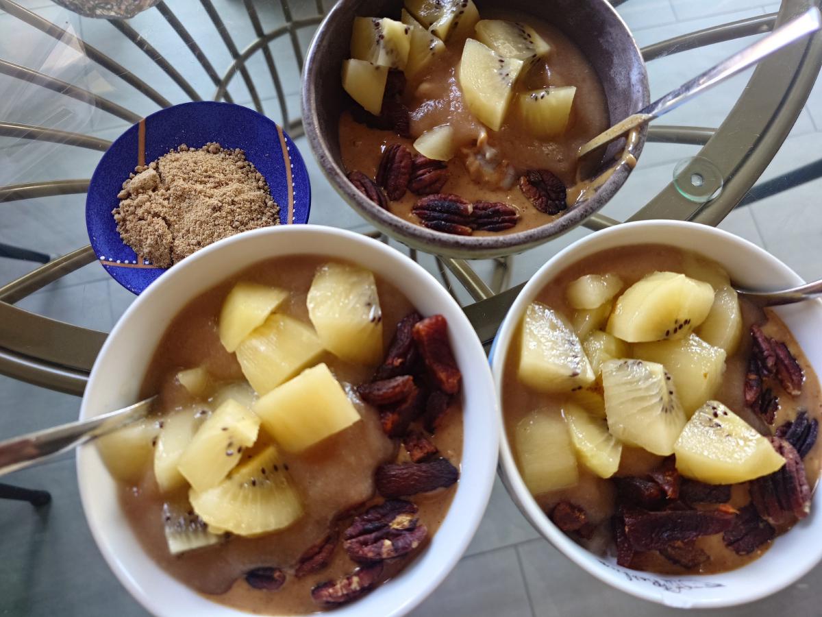 Three bowls of oatmeal topped with gold kiwi, pecans and apricots, a small dish of brown sugar beside them
