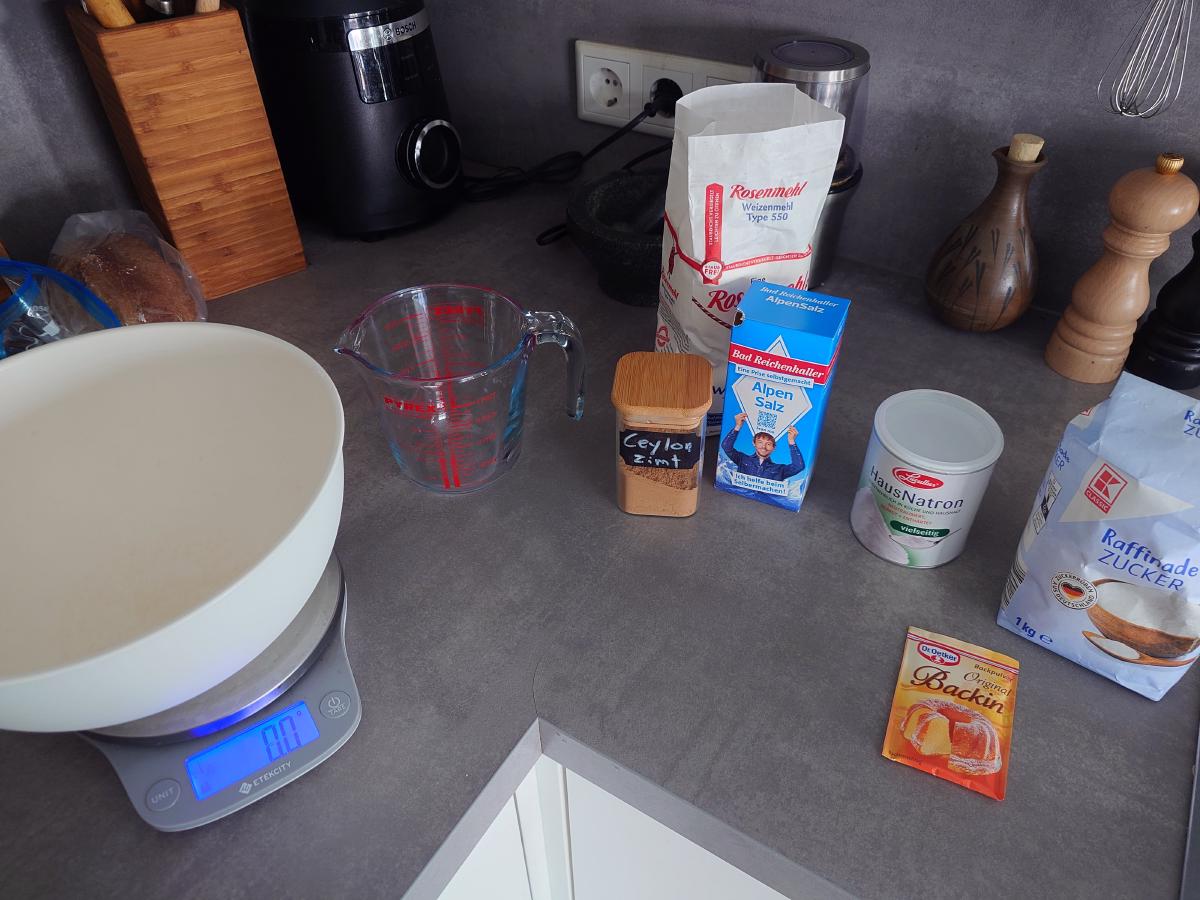 Bowl on the scale with Type 550 flour, sugar, salt, baking soda, baking powder, cinnamon and a measuring jug lined up behind