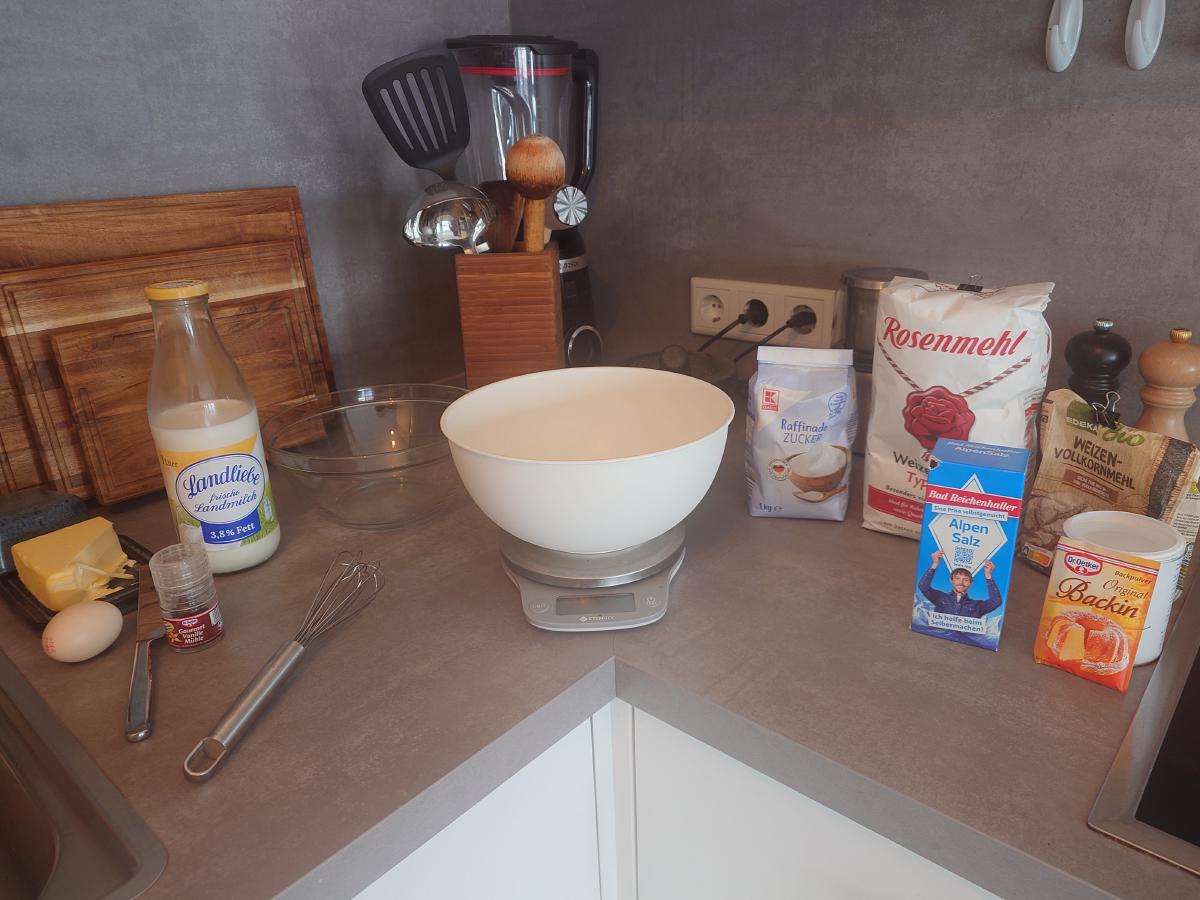 Bowl on the scale with milk, butter, egg, vanilla, flour and sugar lined up behind