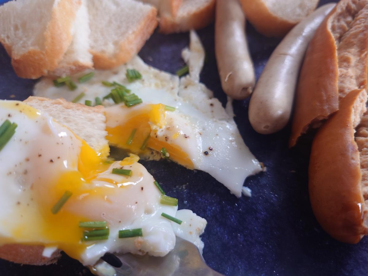 A fried egg cut open, the yolk running across the plate next to bread and sausages
