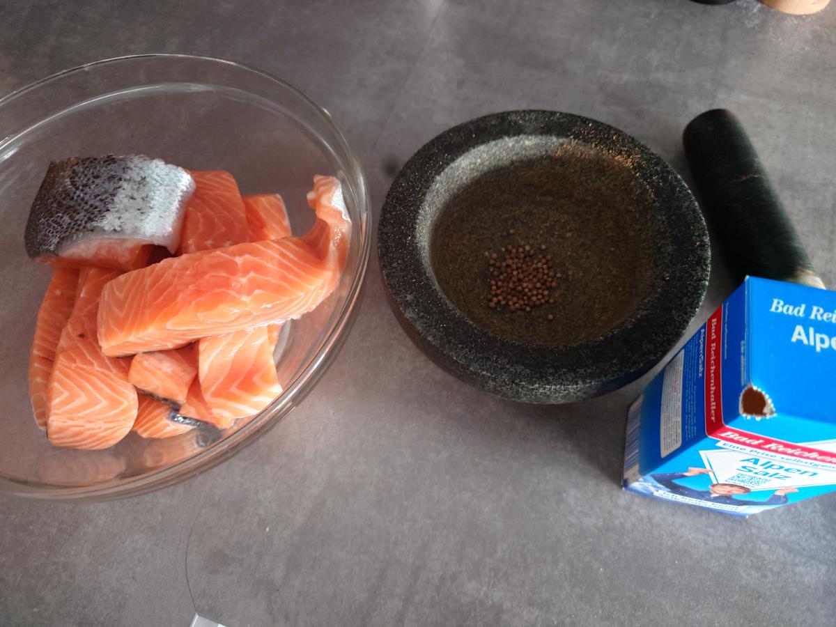 Salmon pieces in a glass bowl beside a granite mortar holding whole coriander seed and a box of salt