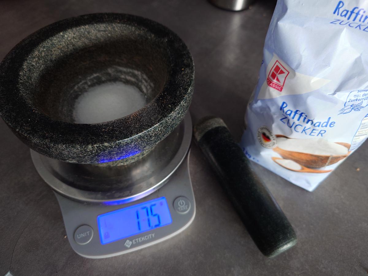 The mortar on the scale reading 17.5 g of sugar, bag of refined sugar beside it