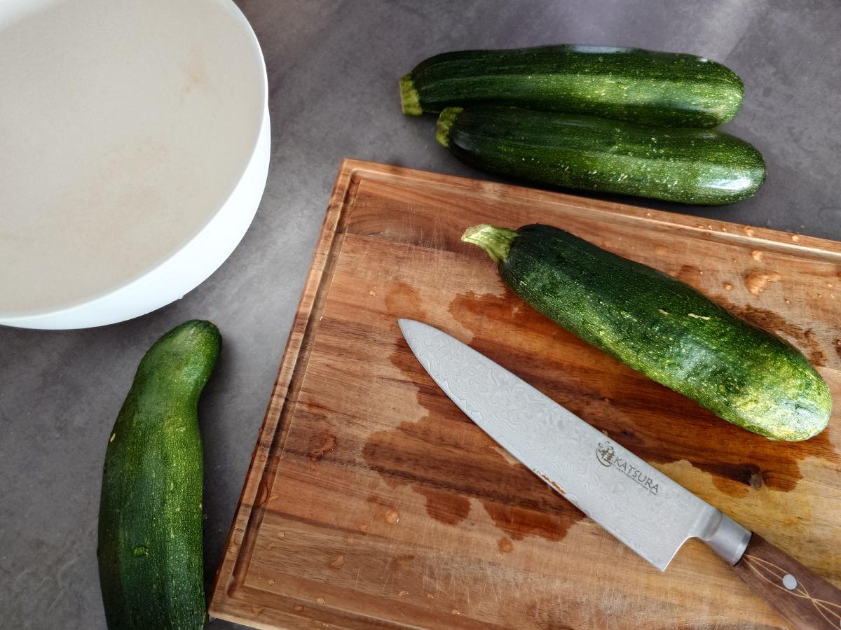 Four zucchini on the counter and board with the knife and an empty white bowl
