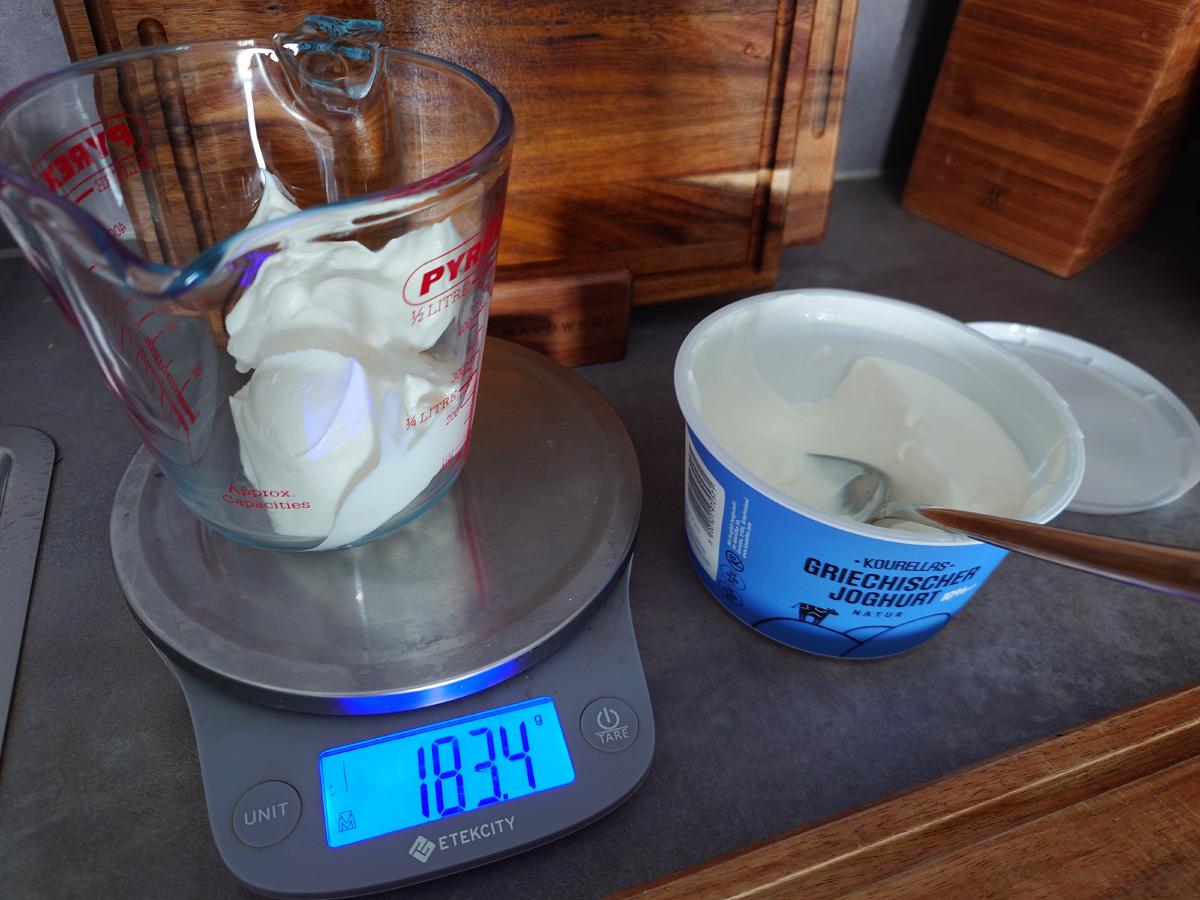 A Pyrex jug of Greek yogurt on the scale reading 183.4 g, tub beside it
